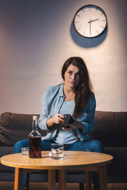 pijana kobieta patrzy w kamerę trzymając pusty portfel przy butelce whisky na stole - Zdjęcie, obraz