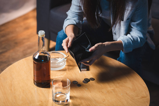 bijgesneden weergave van vrouw met lege portemonnee in de buurt van fles whisky en munten op tafel - Foto, afbeelding