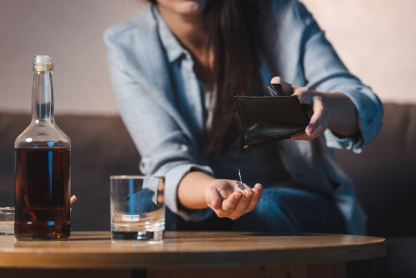 vista parziale della donna che prende monete dal portafoglio vicino bottiglia di whisky sul tavolo, sfondo sfocato - Foto, immagini
