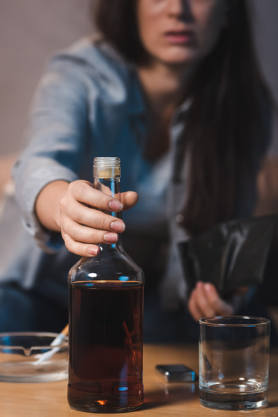 partial view of alcoholic woman taking bottle of whiskey from table, blurred background - Fotoğraf, Görsel