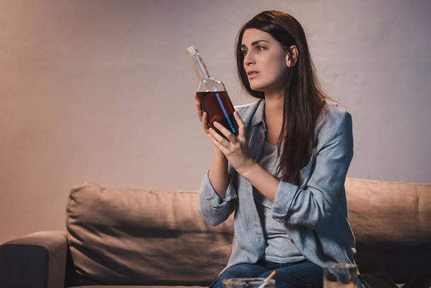 Frustrierte Frau hält Flasche Whiskey in der Hand, während sie allein zu Hause sitzt - Foto, Bild