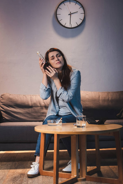 rozrušená, opilá žena držící láhev vína u hlavy, zatímco sedí sama doma - Fotografie, Obrázek