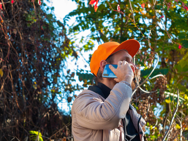 Νεαρός με κόκκινα γένια μιλάει στο κινητό του. Ένας τύπος με αδιάβροχο και πορτοκαλί καπέλο σε πολύχρωμο φόντο φθινοπώρου. Θετικός συναισθηματικός επιχειρηματίας απαντά σε κλήση smartphone. Επικοινωνία τρόπου ζωής. - Φωτογραφία, εικόνα