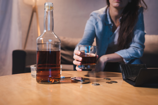 vista parcial de una mujer adicta al alcohol sosteniendo un vaso de whisky cerca de la botella, monedas y billetera vacía, fondo borroso - Foto, Imagen