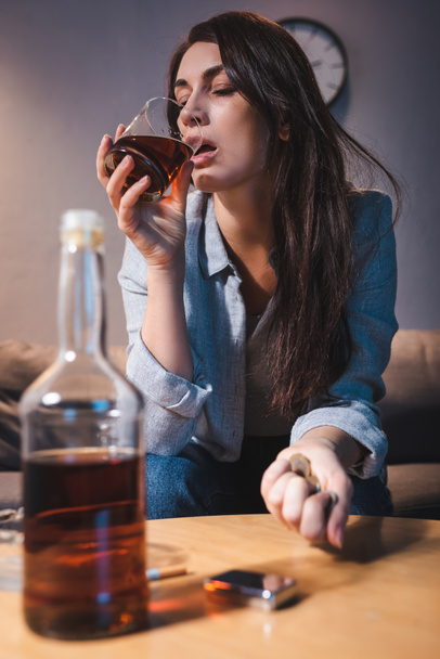 lonely woman drinking whiskey while holding coins near bottle on blurred foreground - Photo, Image