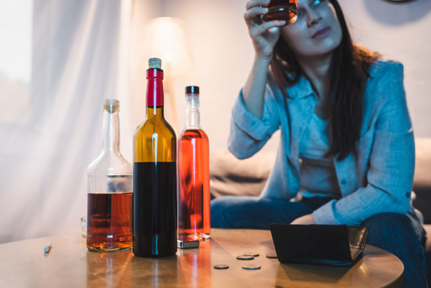 mujer borracha sosteniendo un vaso de alcohol cerca de botellas, monedas y billetera vacía en la mesa, fondo borroso - Foto, Imagen