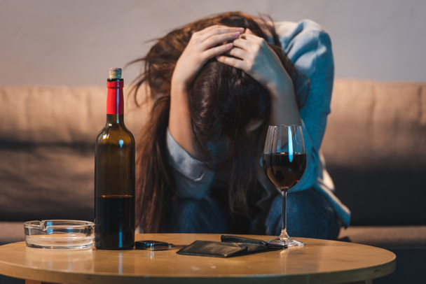 drunk, depressed woman sitting with bowed head near bottle of red wine and empty wallet, blurred background  - Фото, зображення