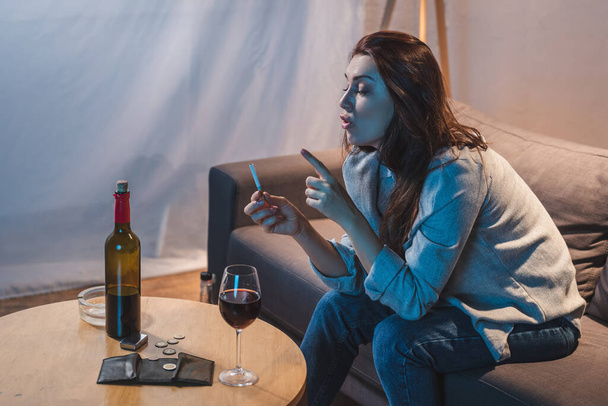 mulher bêbada segurando cigarro enquanto sentado perto de vinho tinto, moedas e carteira vazia na mesa   - Foto, Imagem