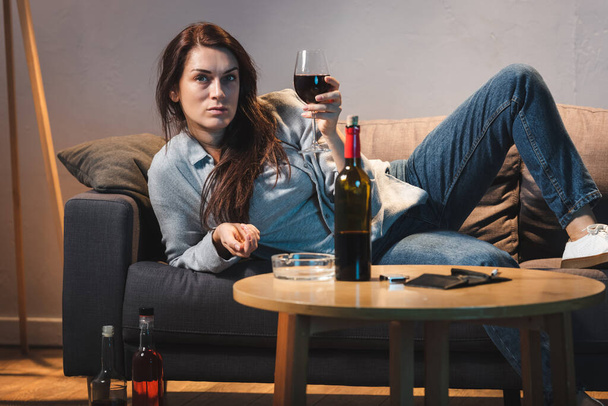 drunk woman with glass of red wine lying on sofa near bottles of alcohol  - Foto, imagen