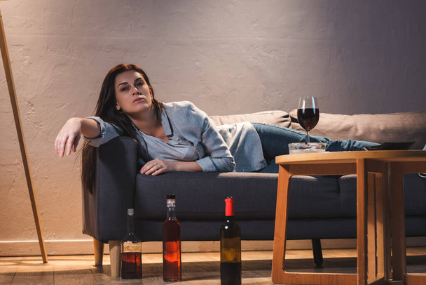 részeg, frusztrált nő a kamerába néz, miközben a kanapén fekszik üvegek mellett a padlón.  - Fotó, kép
