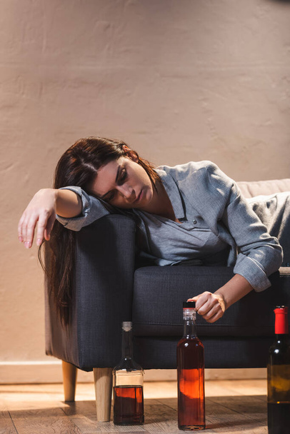 расстроенная пьяная женщина лежит на диване рядом с бутылками на полу - Фото, изображение