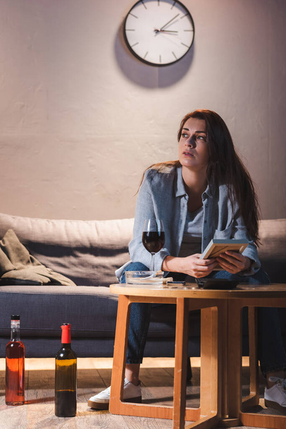 подавленная женщина смотрит в сторону, сидя рядом с бутылками алкоголя и держа фоторамку - Фото, изображение