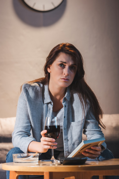 mujer deprimida mirando a la cámara mientras está sentada con un vaso de vino tinto y sosteniendo el marco de la foto    - Foto, imagen