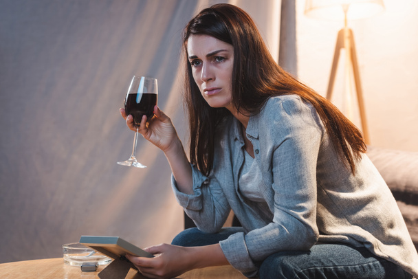 mujer frustrada sosteniendo copa de vino tinto y marco de fotos en casa solo - Foto, Imagen