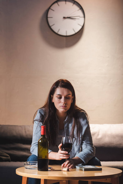 подавленная женщина с бокалом красного вина, сидя в одиночестве возле бутылки и фоторамки на столе - Фото, изображение