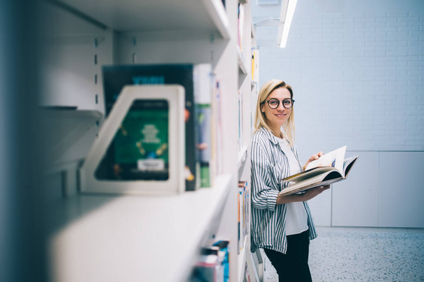 Portrait of successful hipster girl standing in school library with textbook for education and looking at camera, woman in eyewear spending free time for reading best seller enjoy literature hobby - Foto, imagen