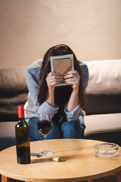 テーブルの上に赤ワインのボトルとガラスの近くに座っている間に写真フレームで顔を隠して落ち込んでいる女性   - 写真・画像