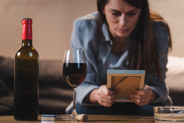 depressive Frau blickt auf Fotorahmen, während sie neben Flasche und Glas Rotwein sitzt, verschwommener Hintergrund    - Foto, Bild