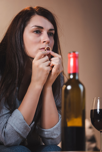 Hayal kırıklığına uğramış bir kadın alyans tutuyor ve bulanık ön planda kırmızı şarap kadehinin yanındaki şişeye bakıyor. - Fotoğraf, Görsel