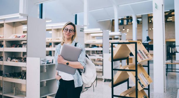 Positive young woman with laptop and hand in pocket looking at camera while standing near bookshelves in modern library in Barcelona City - Zdjęcie, obraz