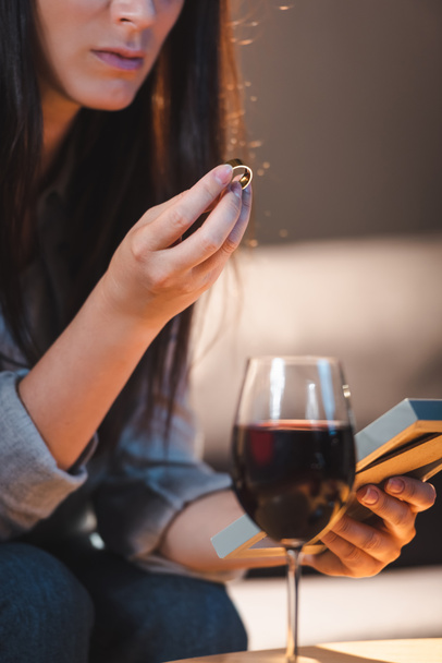 обрезанный вид женщины, держащей фоторамку и обручальное кольцо возле бокала красного вина на размытом переднем плане - Фото, изображение