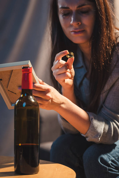 подавленная женщина держит фоторамку и обручальное кольцо возле бутылки красного вина - Фото, изображение