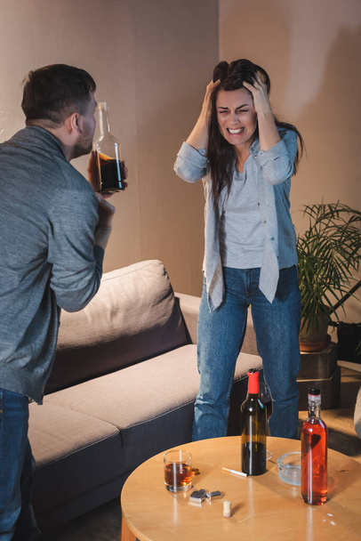 καταθλιπτική, μεθυσμένη γυναίκα αγγίζει το κεφάλι ενώ στέκεται με κλειστά μάτια κοντά στον σύζυγο με ένα μπουκάλι ουίσκι - Φωτογραφία, εικόνα