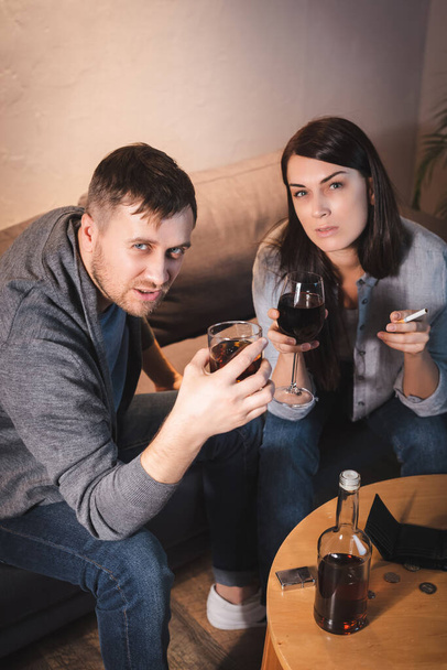 alkoholem závislý pár se dívá na kameru, zatímco drží sklenice whisky a vína - Fotografie, Obrázek