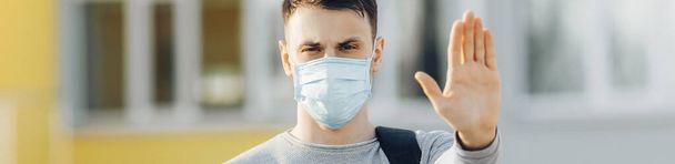 A young man in a protective medical mask on his face, looking at the camera in the open air, shows a palm, a hand, a Stop sign. The concept of the Chinese pandemic of the virus - Valokuva, kuva