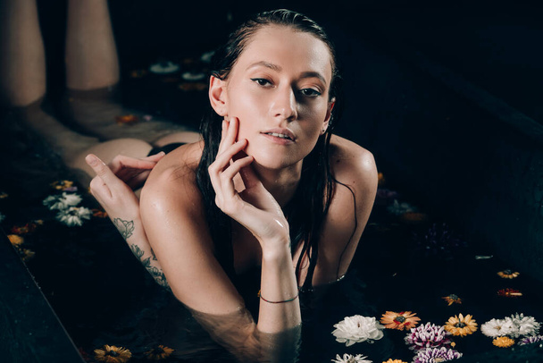Young pretty brunette woman with tattoos in bath with water and flowers. Naked model during an unusual photo shoot in a black bath. - Foto, Imagen