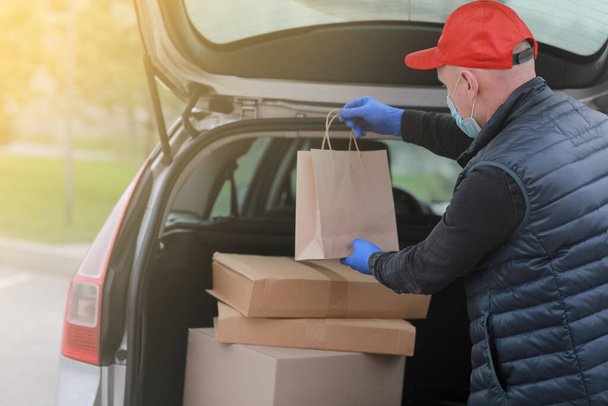 Zusteller mit roter Mütze, Mundschutz und Handschuhen halten Papiertüte in der Nähe des Autos. Service Coronavirus. Online-Shopping. Attrappe - Foto, Bild