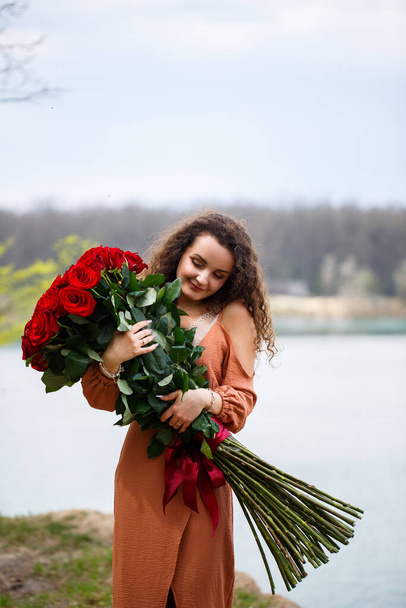 A beautiful girl of European appearance with curly hair and a smile on her face with a huge bouquet of red roses on a background of blue lake. Warm summer day, happy young woman, emotions of joy - Photo, image