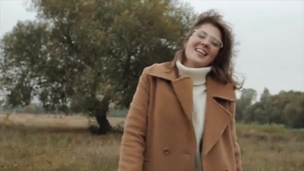 Energická mladá dívka v brýlích stojí na pozadí stromu a směje se, zatímco se zabalí do teplého bílého svetru a kabátu - Záběry, video