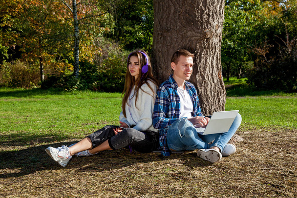 A young couple spends time together in the park, studies and works while sitting near a tree with a laptop, headphones and a phone. Guy with a girl with gadgets in the park - Fotografie, Obrázek