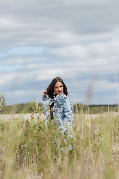 Aantrekkelijk vrouwelijk model in jeans kijkend naar camera en borst bedekkend met denim jas op wazige achtergrond van het platteland - Foto, afbeelding