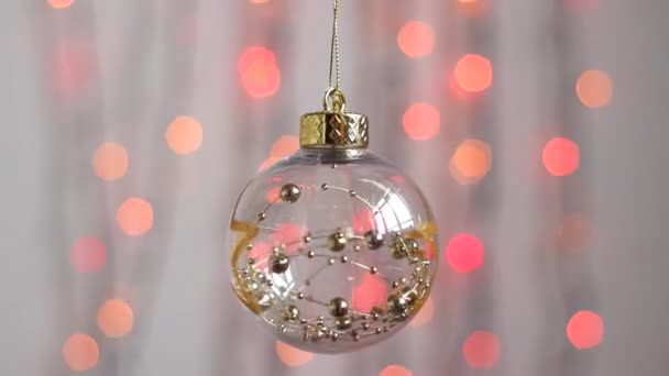 Krásné vánoční stromeček hračka visí na pozadí multi-barevné vánoční světla a pohybuje mírně. Detailní záběr. Průhledná vánoční stromeček hračka v podobě koule se zlatými hvězdami a bal - Záběry, video