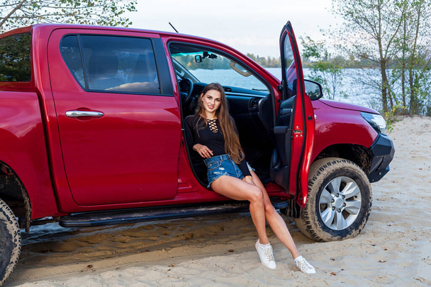 Beautiful sexy girl with long hair in short denim shorts and a black bodysuit posing near the open door of a red pickup truck. Girl posing near the car - Foto, imagen