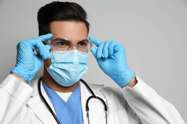 Doctor in protective mask, glasses and medical gloves against light grey background - 写真・画像