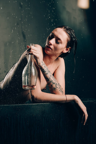 Joven mujer morena bonita con tatuajes en un baño. Modelo desnudo durante una sesión de fotos inusual en un baño negro beber vino - Foto, imagen