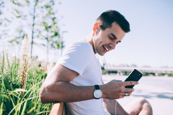 Zijaanzicht van een glimlachende jonge man die alleen op de bank zit in het stadspark in de buurt van planten die oortelefoons en smartphones gebruiken om via videogesprekken te praten - Foto, afbeelding