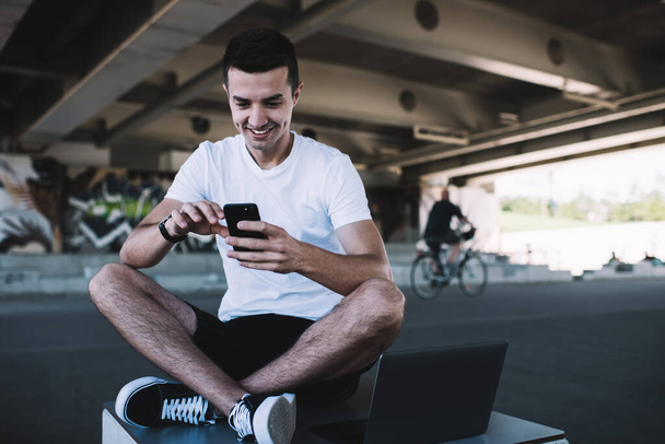 Happy youthful male in informal wear chatting with friend on mobile phone while smiling and sitting under bridge with crossed legs - Foto, Bild