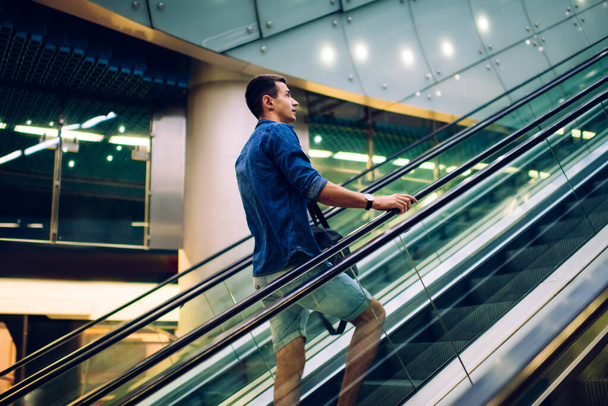 Low angle side view of young male in casual outfit going up on escalator at modern building with glass walls - Fotoğraf, Görsel