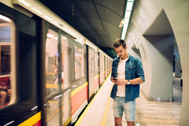Serious young guy in denim standing near metro train on platform of subway station while browsing cellphone in soft light - 写真・画像