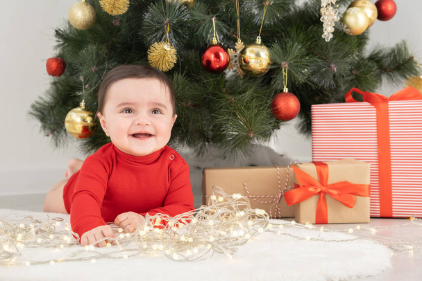 baby girl in red bodysuit under Christmas tree with gift boxes presents. warm winter evening at home - Photo, Image