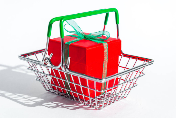 Small grocery basket with red gift box on white isolated background. Creative Christmas shopping online. Holiday sales and discounts on new year and black Friday. Trading business. Hard shadows. - Fotografie, Obrázek