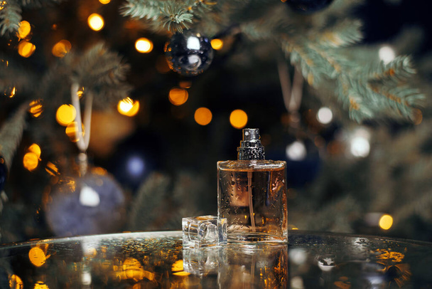 Beautiful composition of a bottle of women's perfume with drops of water on a glass table against the background of a Christmas tree. New year gift, garland lights, toys and holiday concept. - Foto, Bild