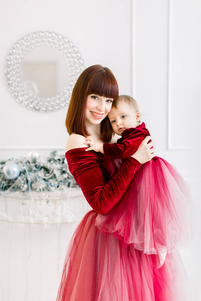 Christmas and Happy New Year Holidays. Family, mother and child concept. Tender photo of happy pretty mom holding and hugging her cute baby girl in red dress, having fun at decorated room - 写真・画像