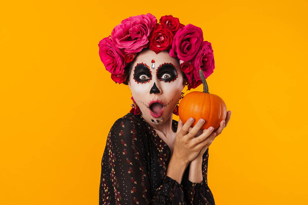 Shocked young girl in halloween makeup posing with pumpkin isolated over yellow background - Zdjęcie, obraz