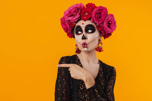 Amusing girl in halloween makeup and flower wreath pointing fingers aside isolated over yellow wall - Foto, Imagen