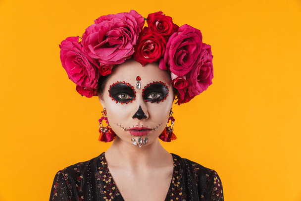 Beautiful girl in halloween makeup and flower wreath posing at camera isolated over yellow wall - Photo, image